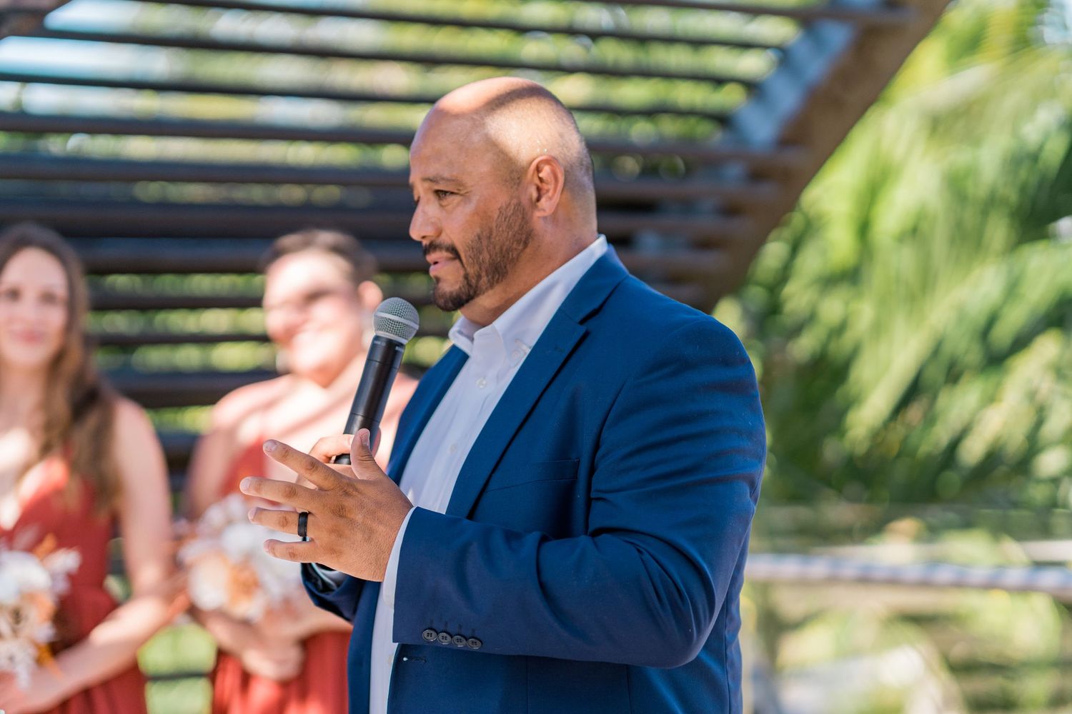Man in blue blazer speaks into microphone at outdoor wedding ceremony with bridal party.