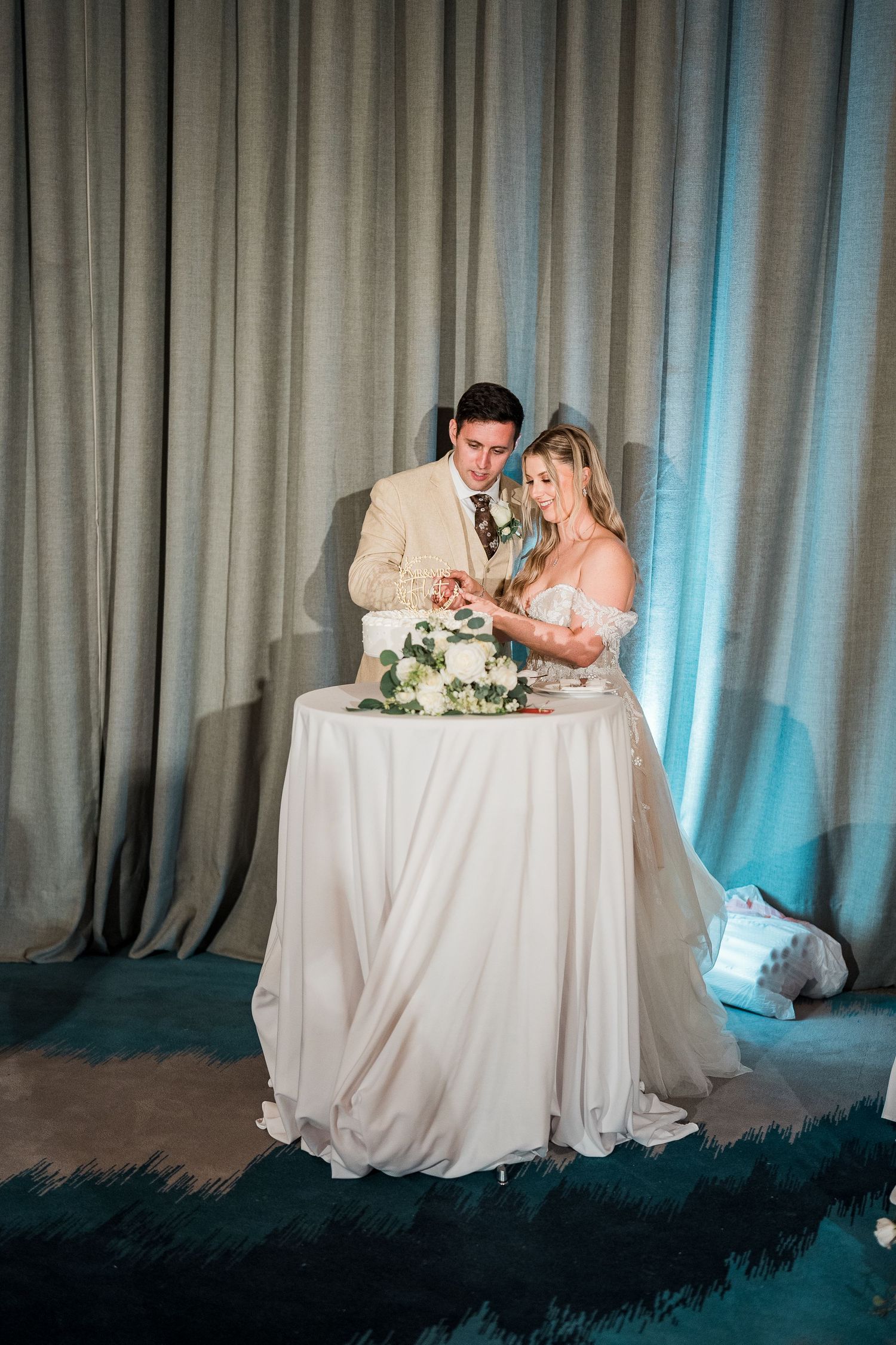 A bride and groom cut their wedding cake together in front of elegant draped curtains indoors.