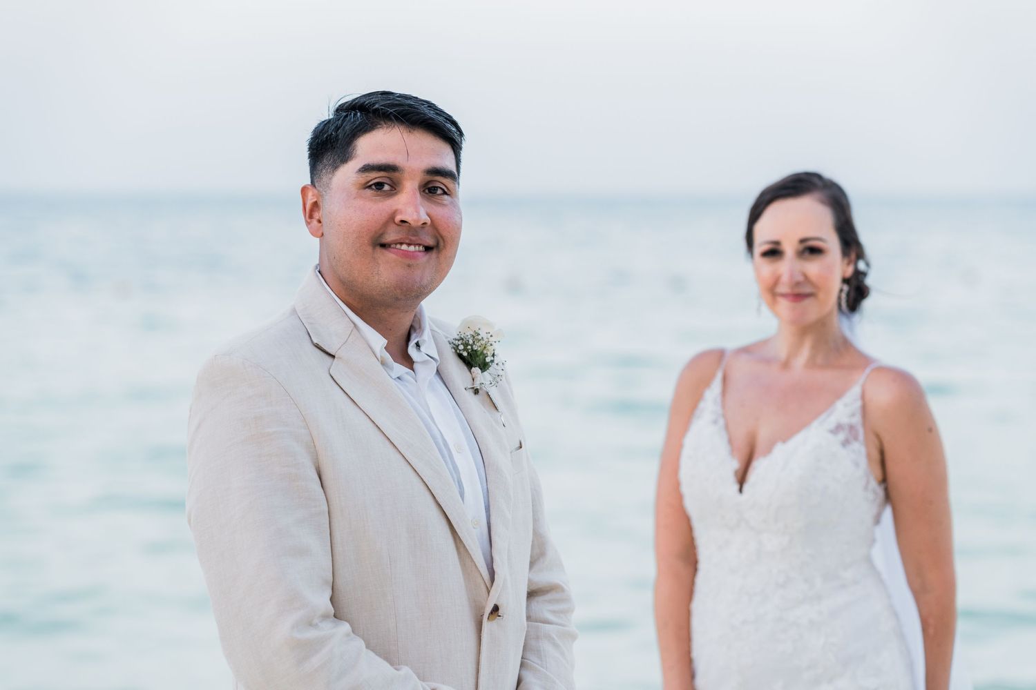 Groom in tan suit smiling with bride in white gown on beach.
