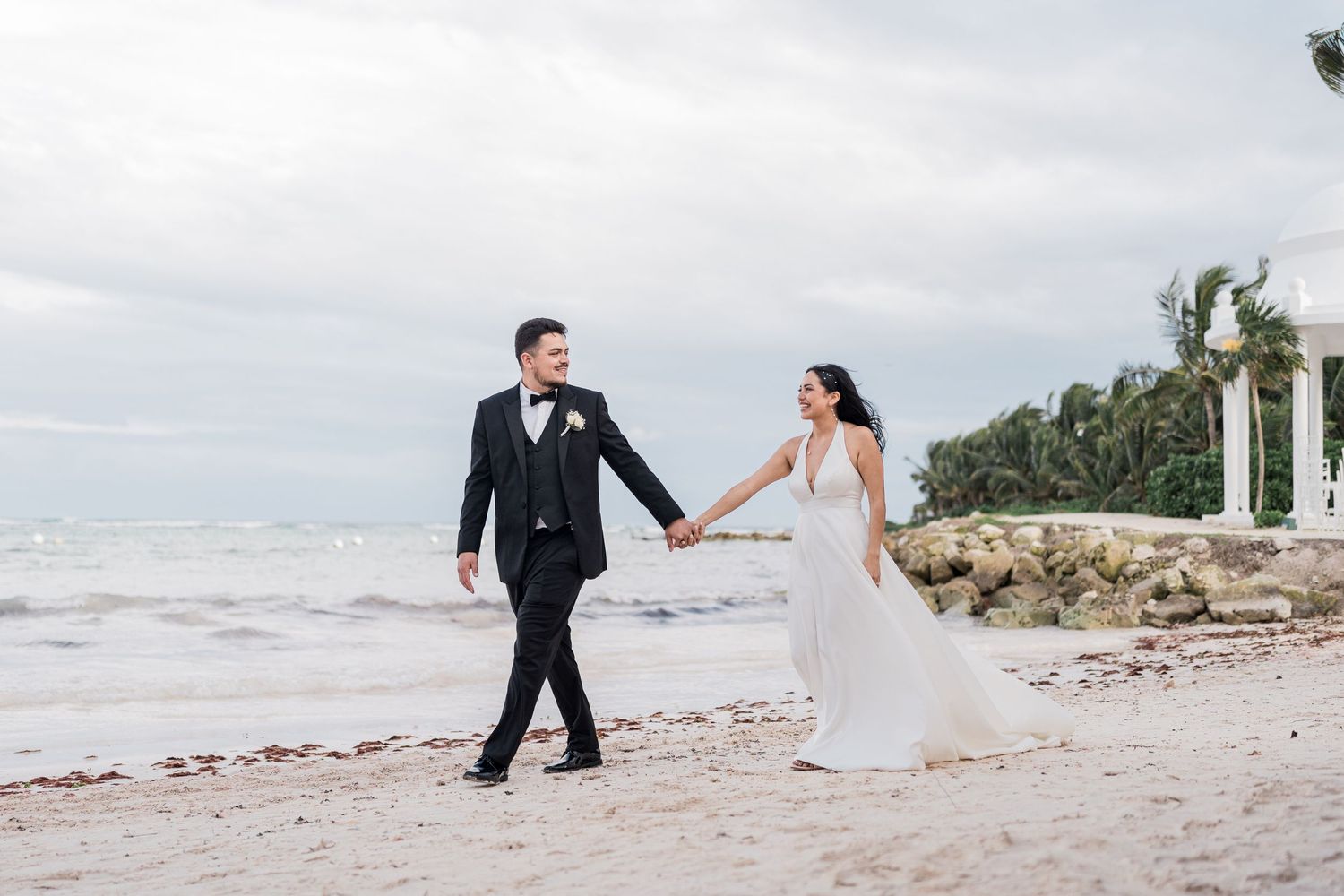 Bride and groom walk hand in hand along a sandy beach with ocean waves and white arches.