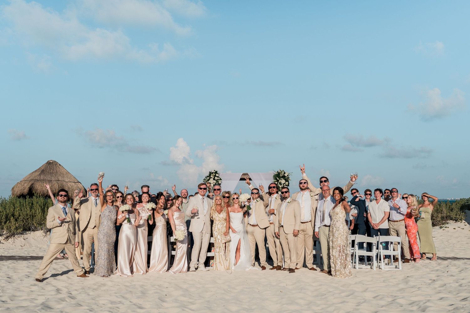 Wedding party group photo on sandy beach with thatched hut backdrop under clear blue sky.