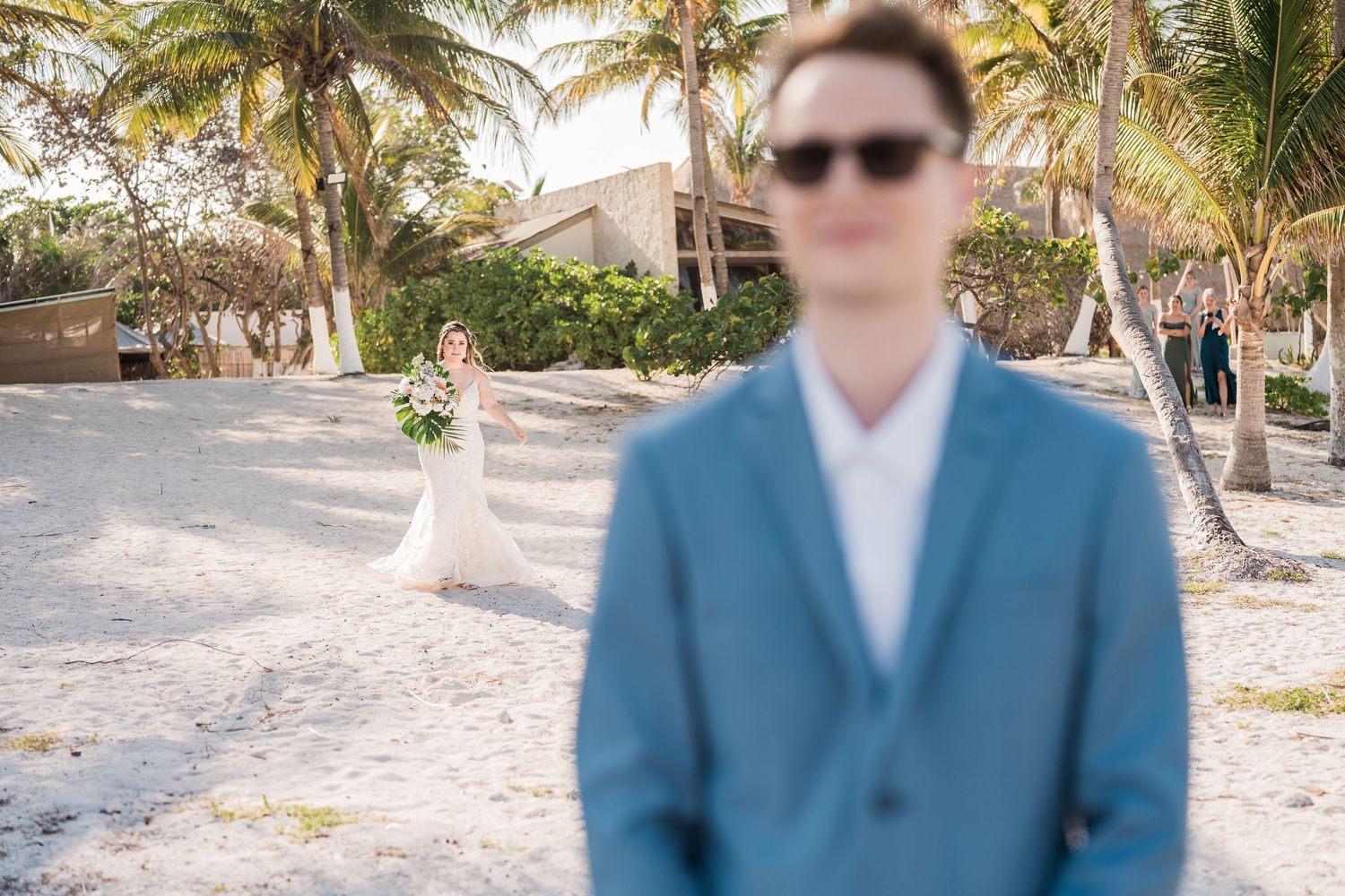 Groom in blue suit with sunglasses awaits bride approaching through palm tree-lined beach path.