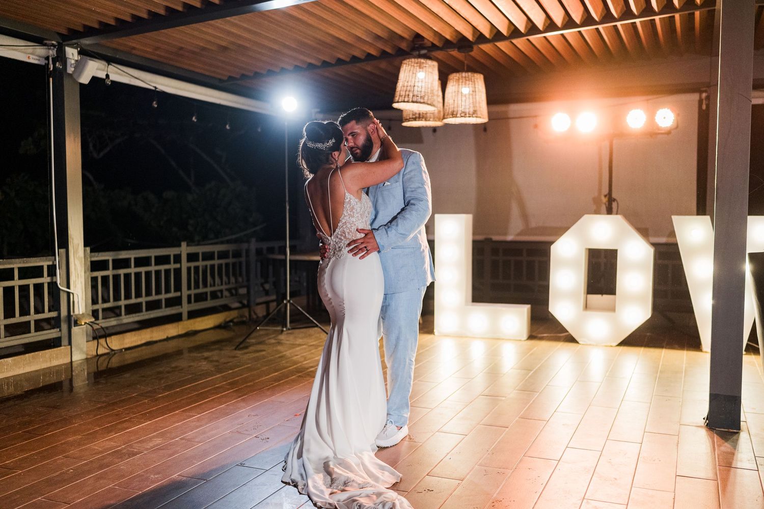 Bride and groom sharing romantic first dance embrace on outdoor terrace with illuminated LOVE marquee letters behind them.