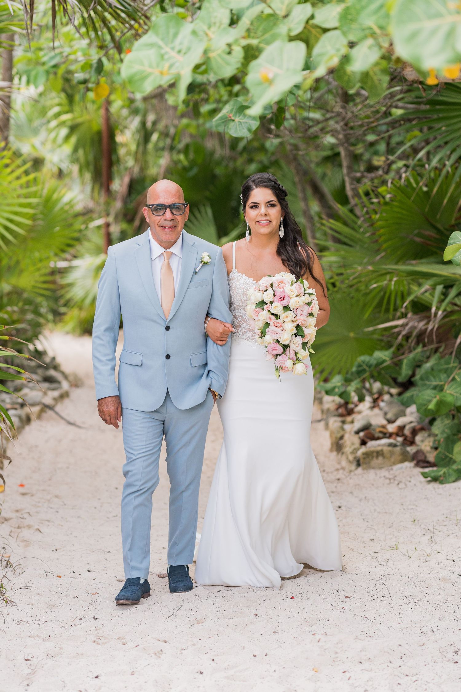 Father of the bride walking his daughter down a sandy tropical path lined with lush green foliage.