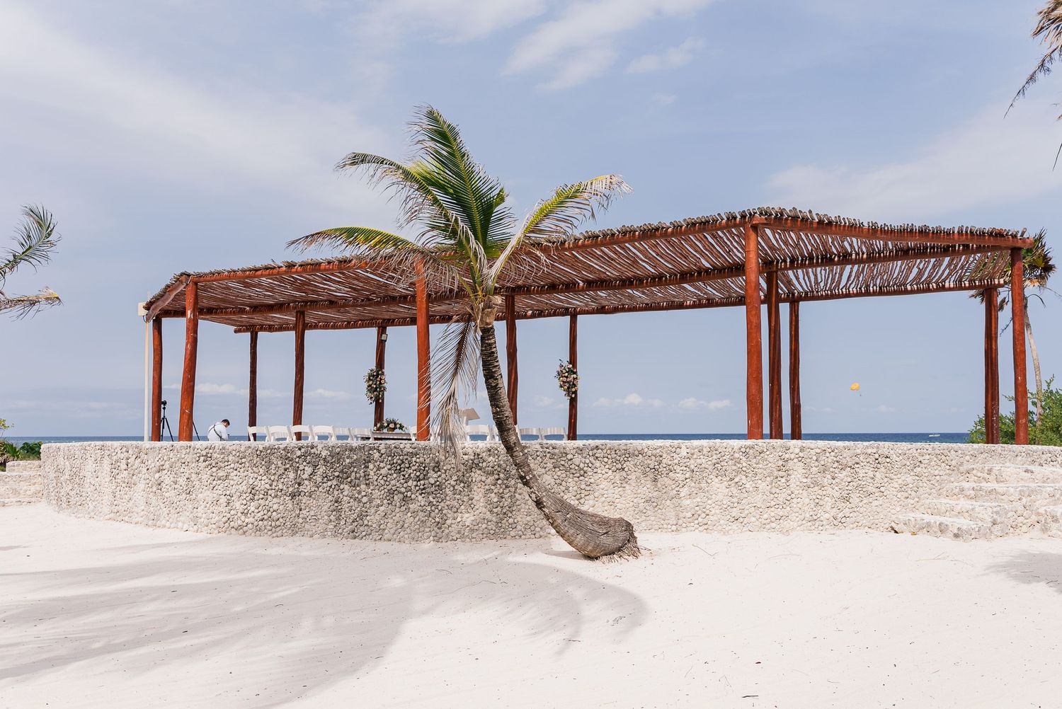 Rustic wooden beach pergola with leaning palm tree on white sand at tropical wedding venue.