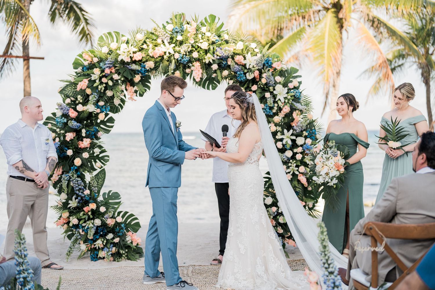 Couple exchanges vows under tropical floral arch with green-dressed bridesmaids at beach wedding.