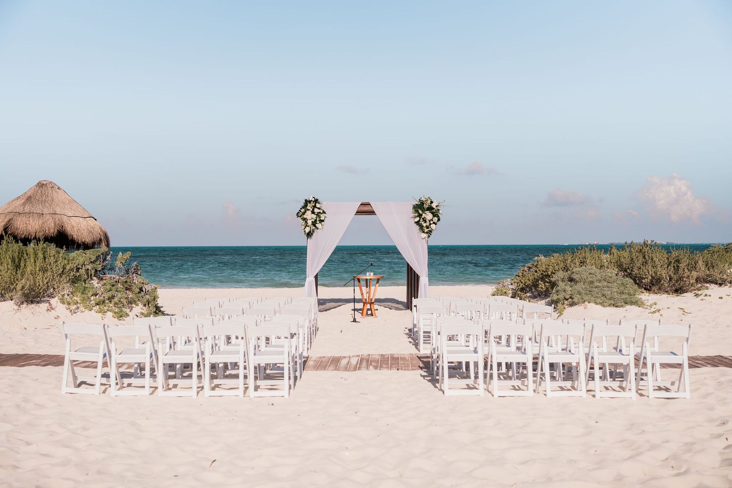 Elegant beach wedding ceremony setup with white chairs and floral arch overlooking turquoise ocean.