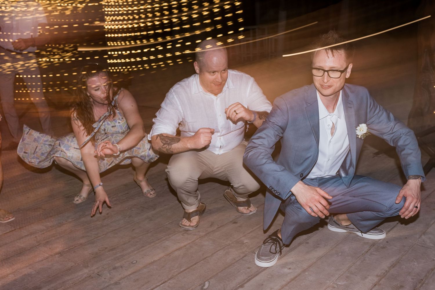 Wedding guests do low squat dance move on wooden dance floor at lively reception.