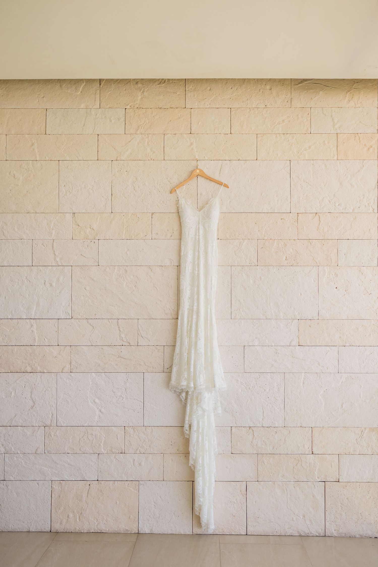 Elegant white wedding dress hanging on a wooden hanger against a beige stone brick wall.