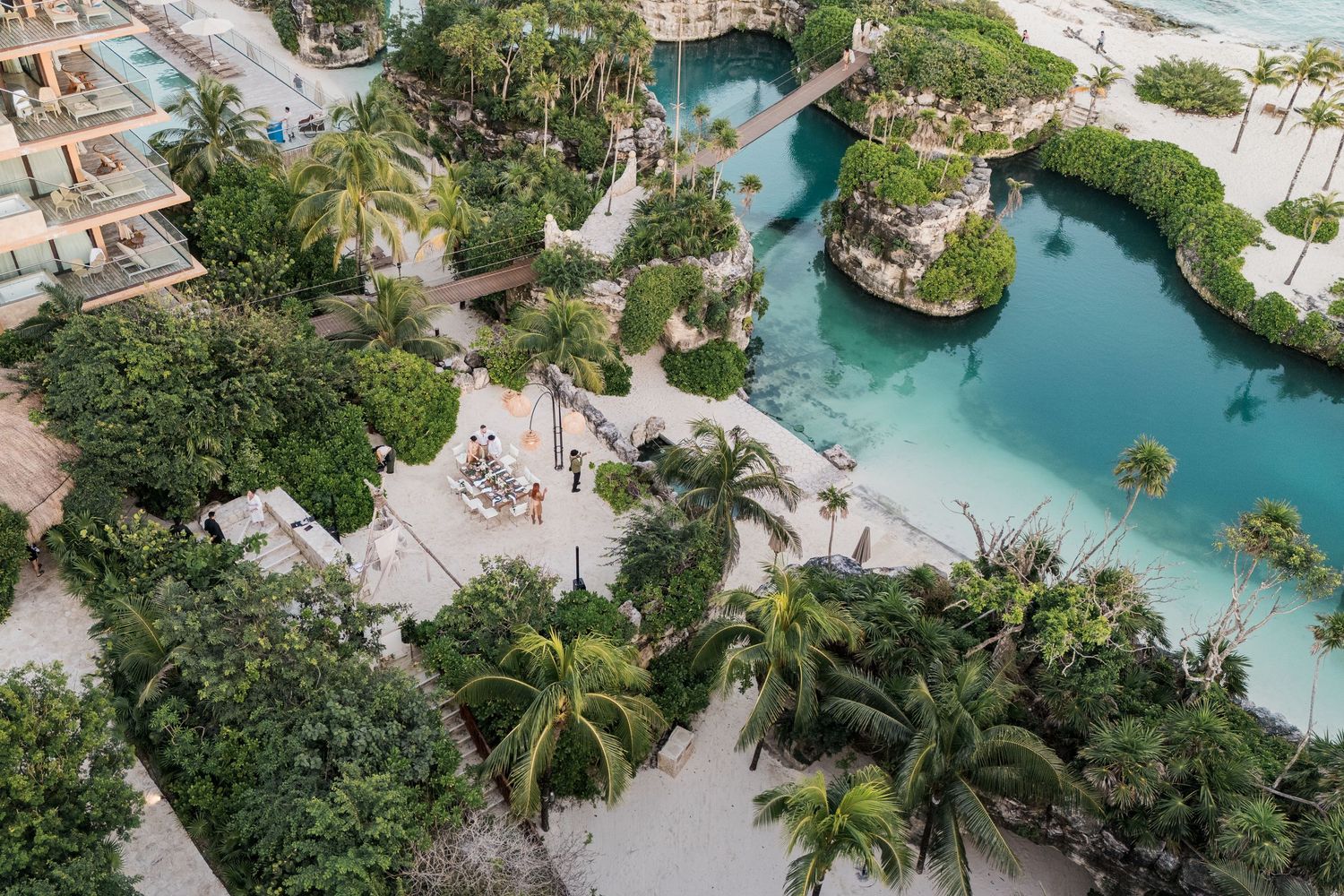 Xcaret Arte Wedding - Aerial view of a tropical resort lagoon surrounded by lush palm trees and sandy pathways with turquoise water.