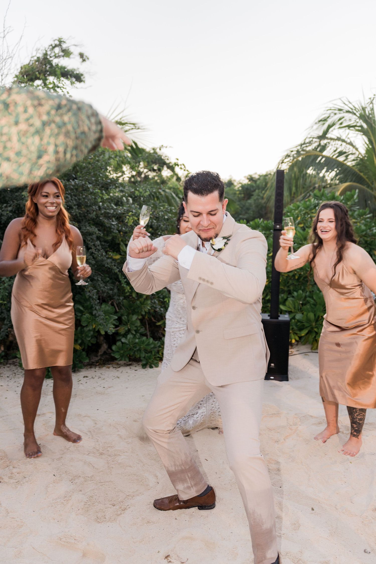 Xcaret Arte Wedding - Groom dancing on sandy beach at wedding reception with bridesmaids in gold dresses holding champagne glasses.