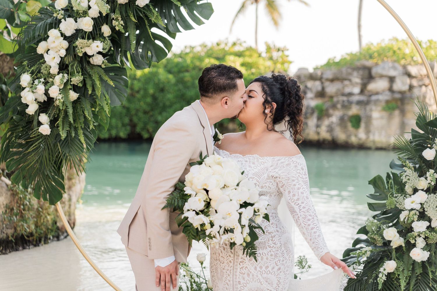 Xcaret Arte Wedding - Bride and groom kissing under floral arch with white roses and tropical greenery at a beachside wedding ceremony.