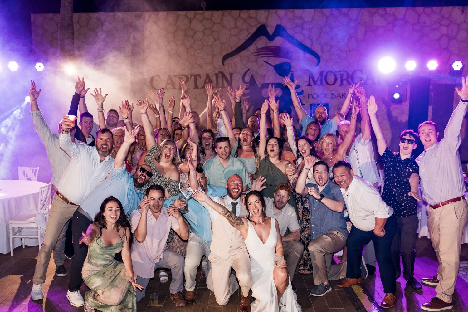 Large wedding party celebrating under Captain Morgan sign with raised arms and big smiles at a festive outdoor reception.