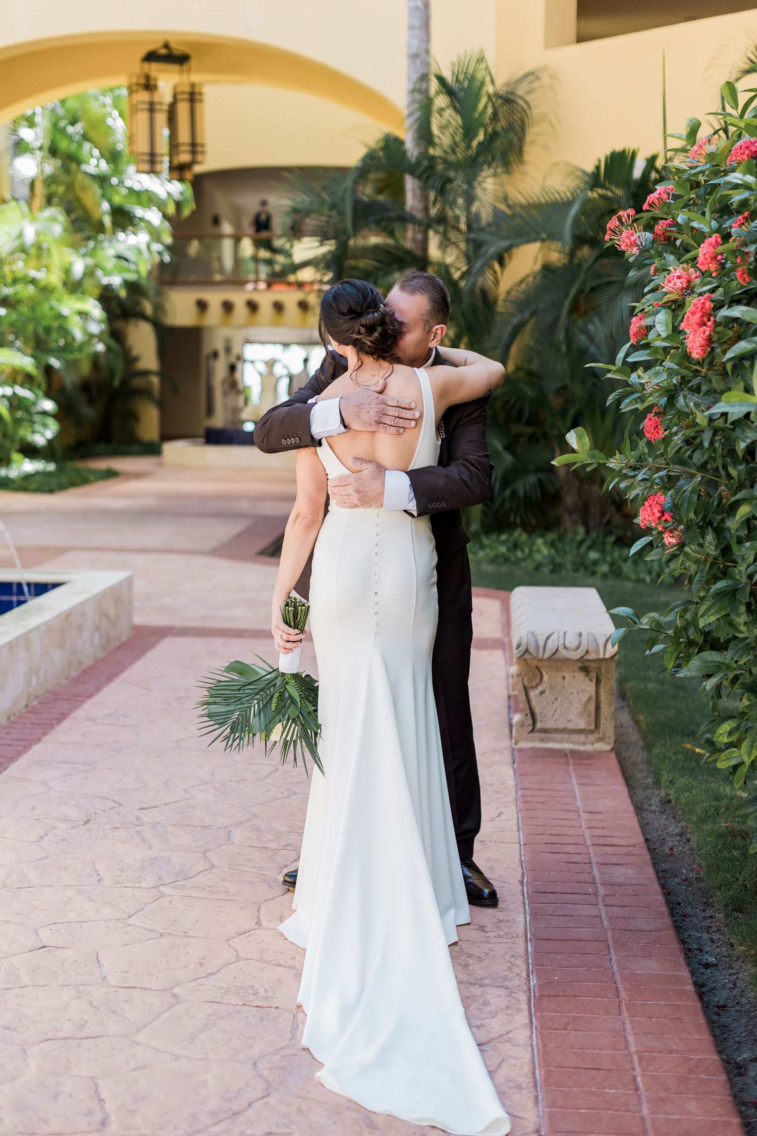 Bride and groom share romantic embrace outdoors at elegant resort venue, bride in sleek white gown holding tropical bouquet.