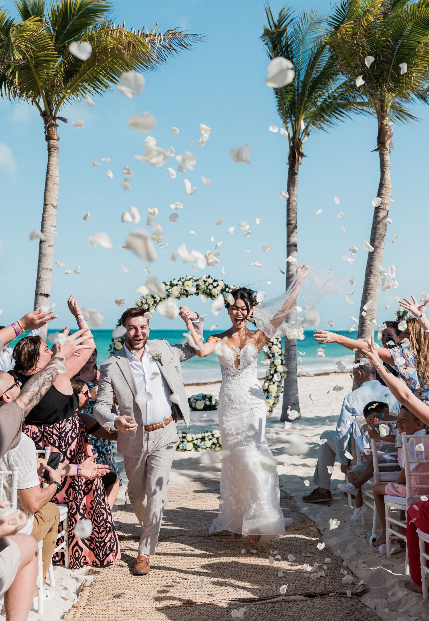 Joyful newlyweds walk down beach aisle as guests toss white flower petals, with palm trees and ocean backdrop.