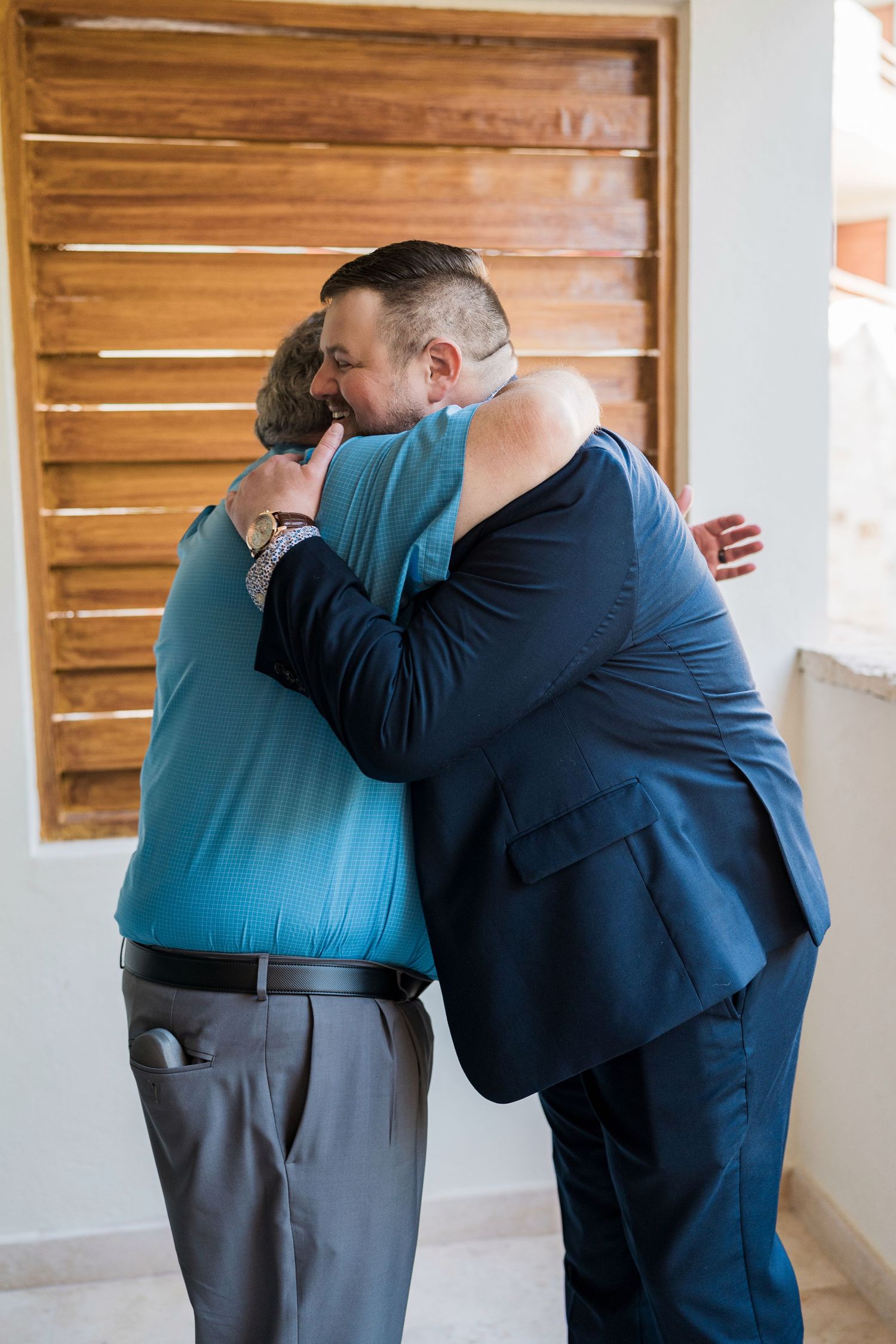 Secrets Akumal Wedding - A man in a navy suit shares a warm embrace with an older woman in a teal top near wooden shutters.
