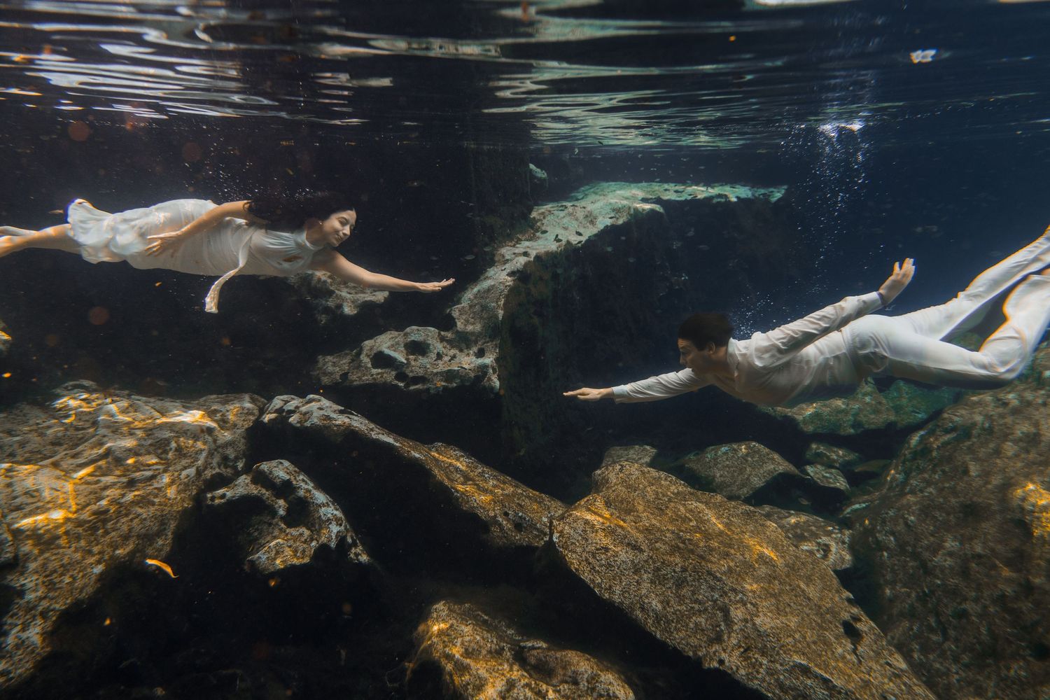 Two women in white dresses swimming gracefully underwater among rocky formations in clear water.