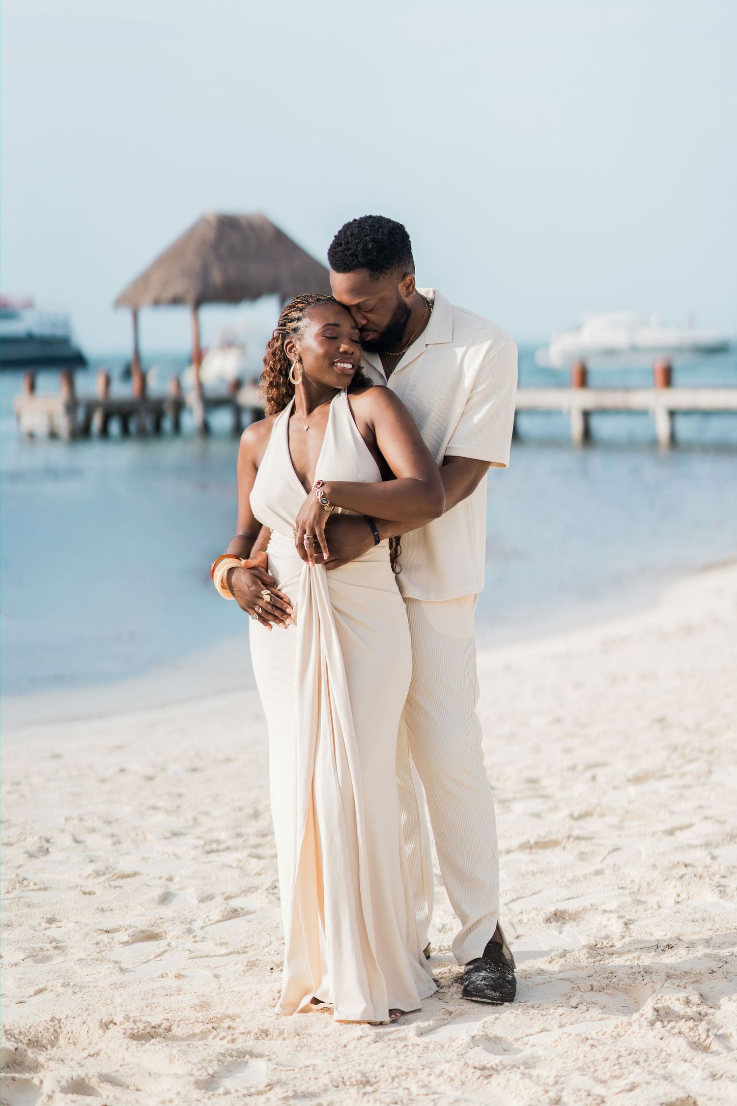 Couple in cream outfits embracing on a white sand beach with a pier and calm blue water in the background.