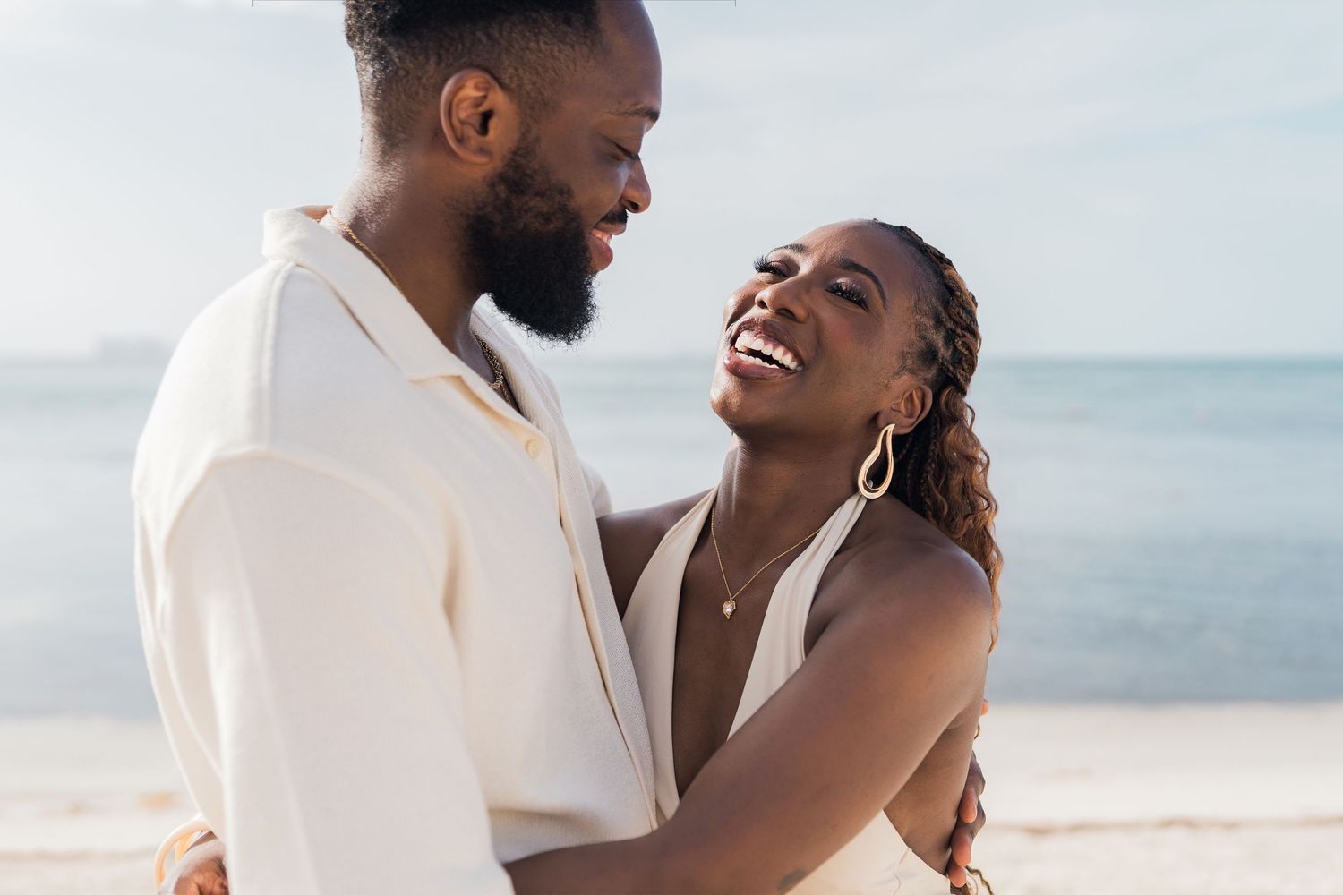 Happy Black couple embracing and smiling on a sunny beach, wearing white outfits with ocean in the background.