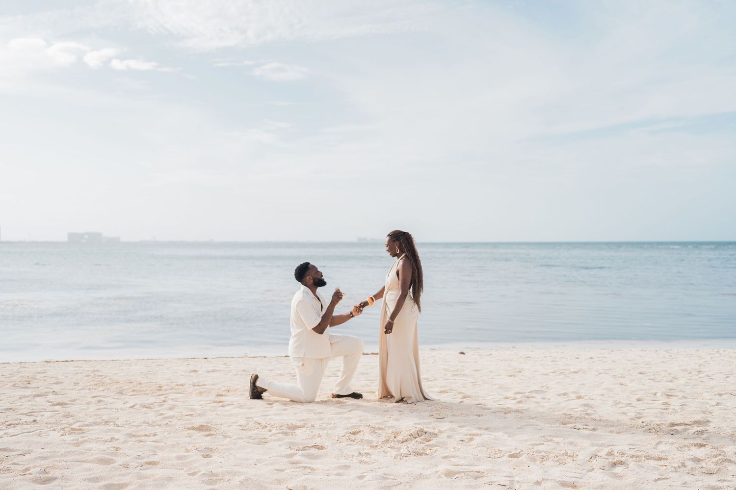 Man kneeling on beach proposing to woman in white dress by the ocean.