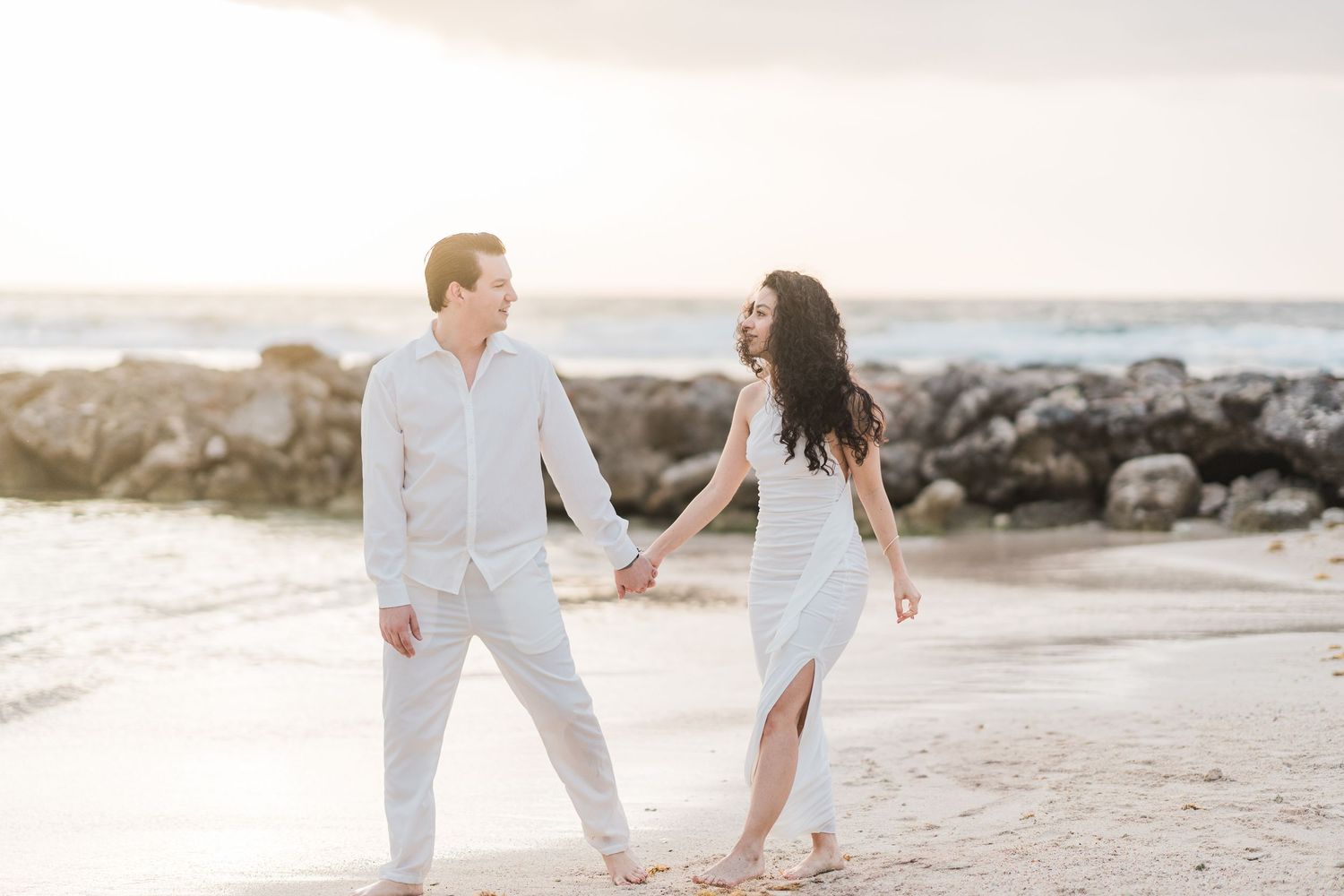 Couple in white outfits holding hands and walking on a sandy beach at sunset with rocky shoreline backdrop.