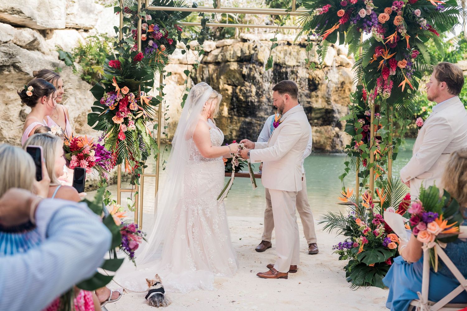 Couple exchanges vows at tropical outdoor wedding ceremony with vibrant floral arch near rocky waterfall.