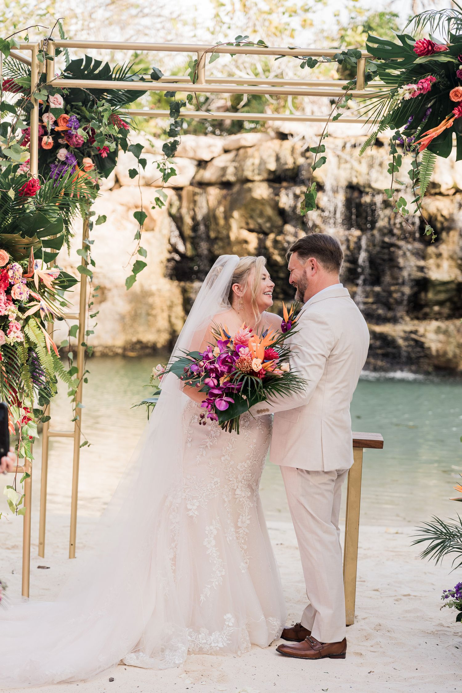 Bride and groom share a joyful moment under a tropical floral arch on a sandy beach during their destination wedding.