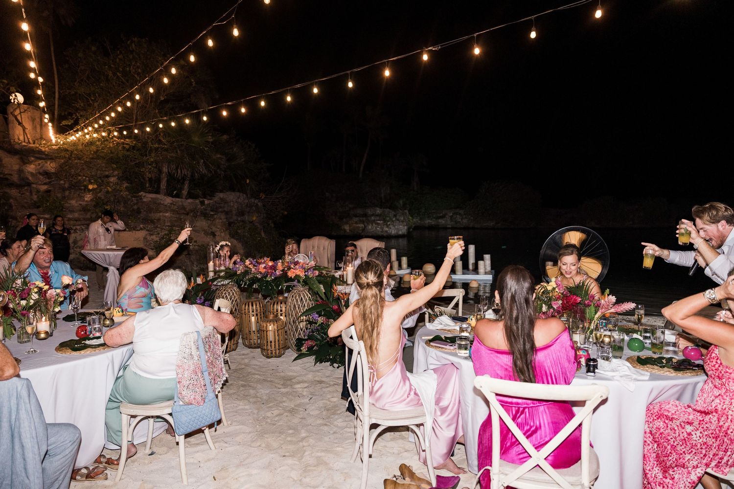 Guests raise glasses in a toast at an outdoor evening wedding reception with string lights and tropical floral centerpieces.