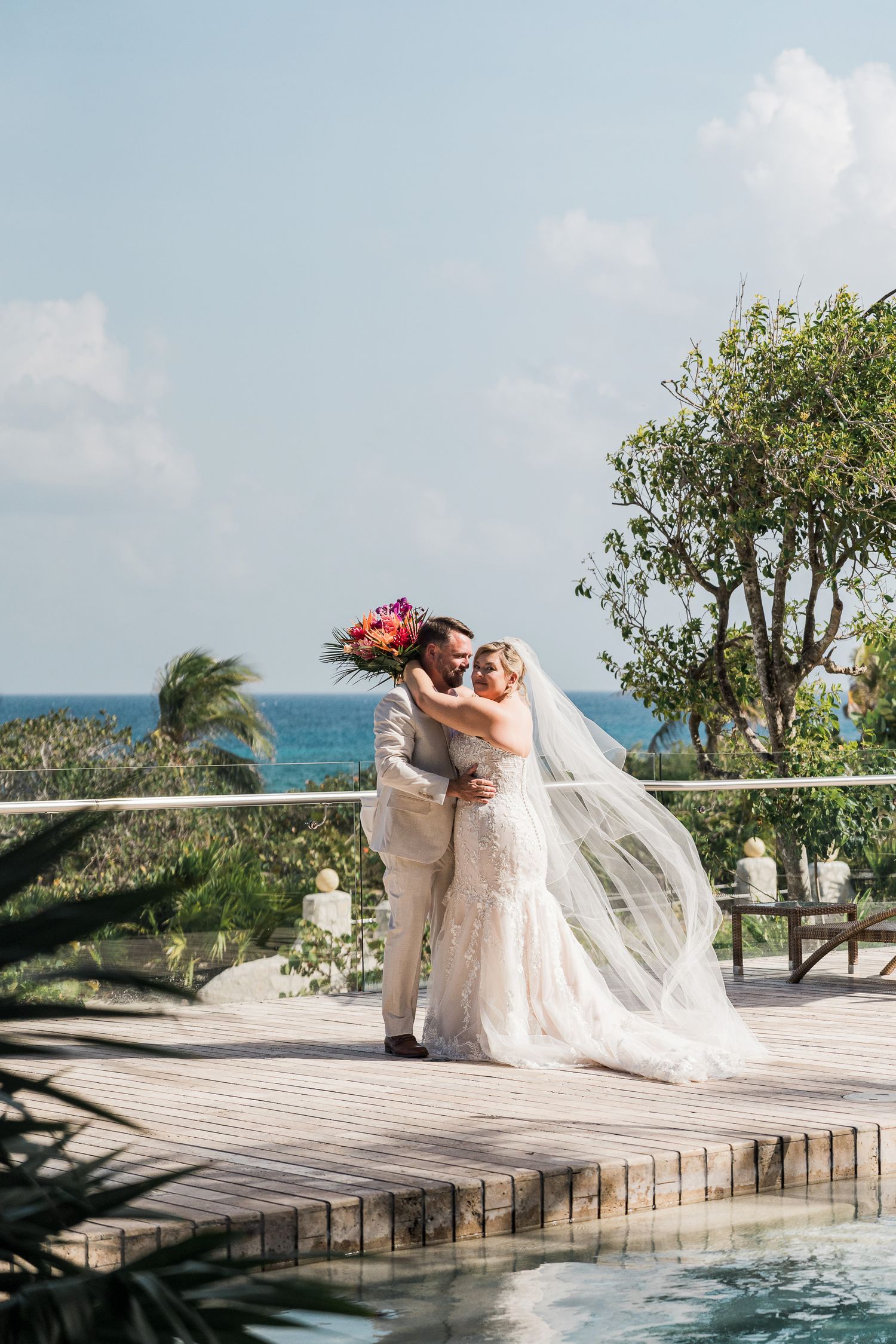 Bride and groom embrace on wooden deck overlooking turquoise ocean, bride in lace gown with flowing veil holding colorful bouquet.
