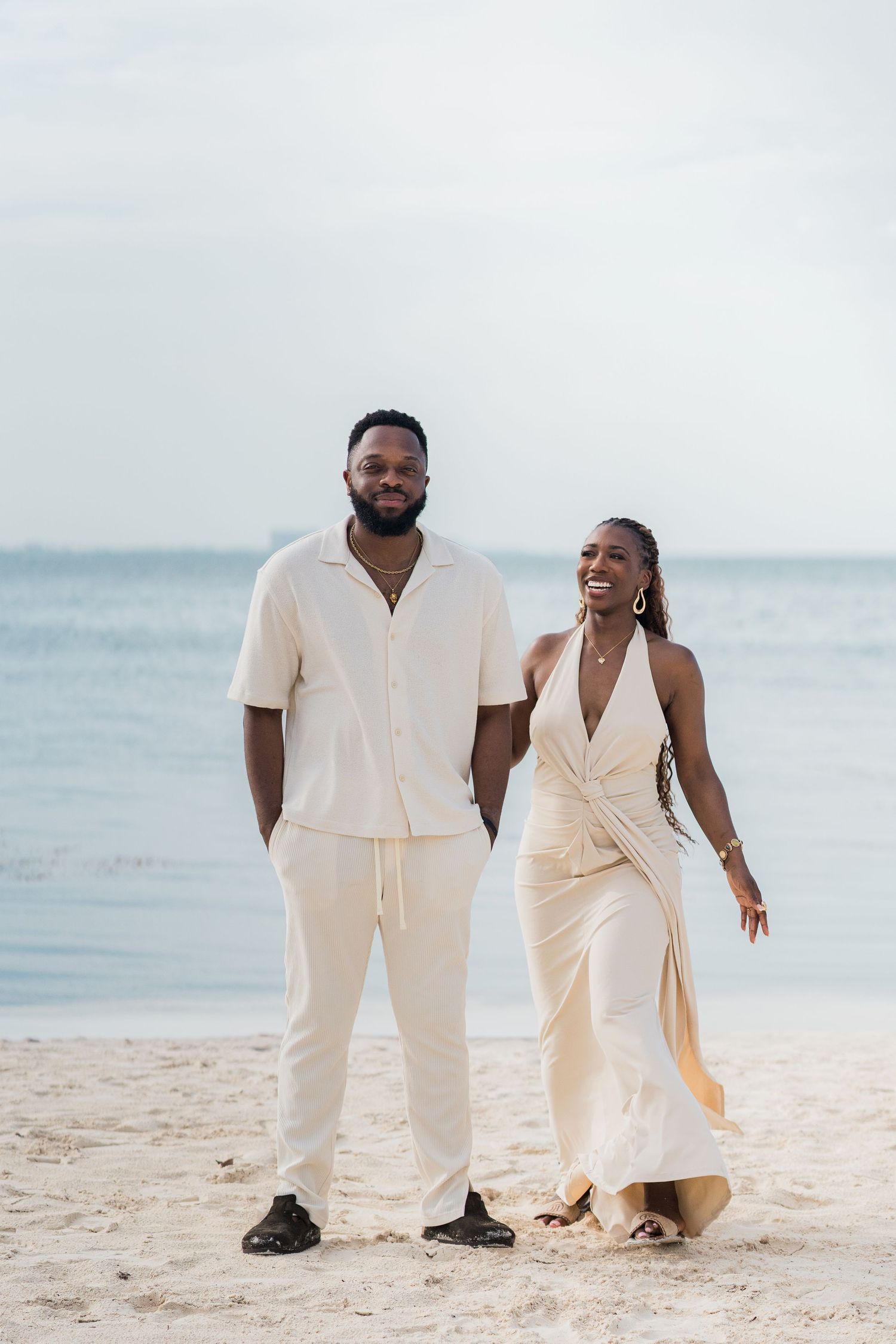 Couple in matching cream outfits walking on a sandy beach with calm ocean waters in the background.
