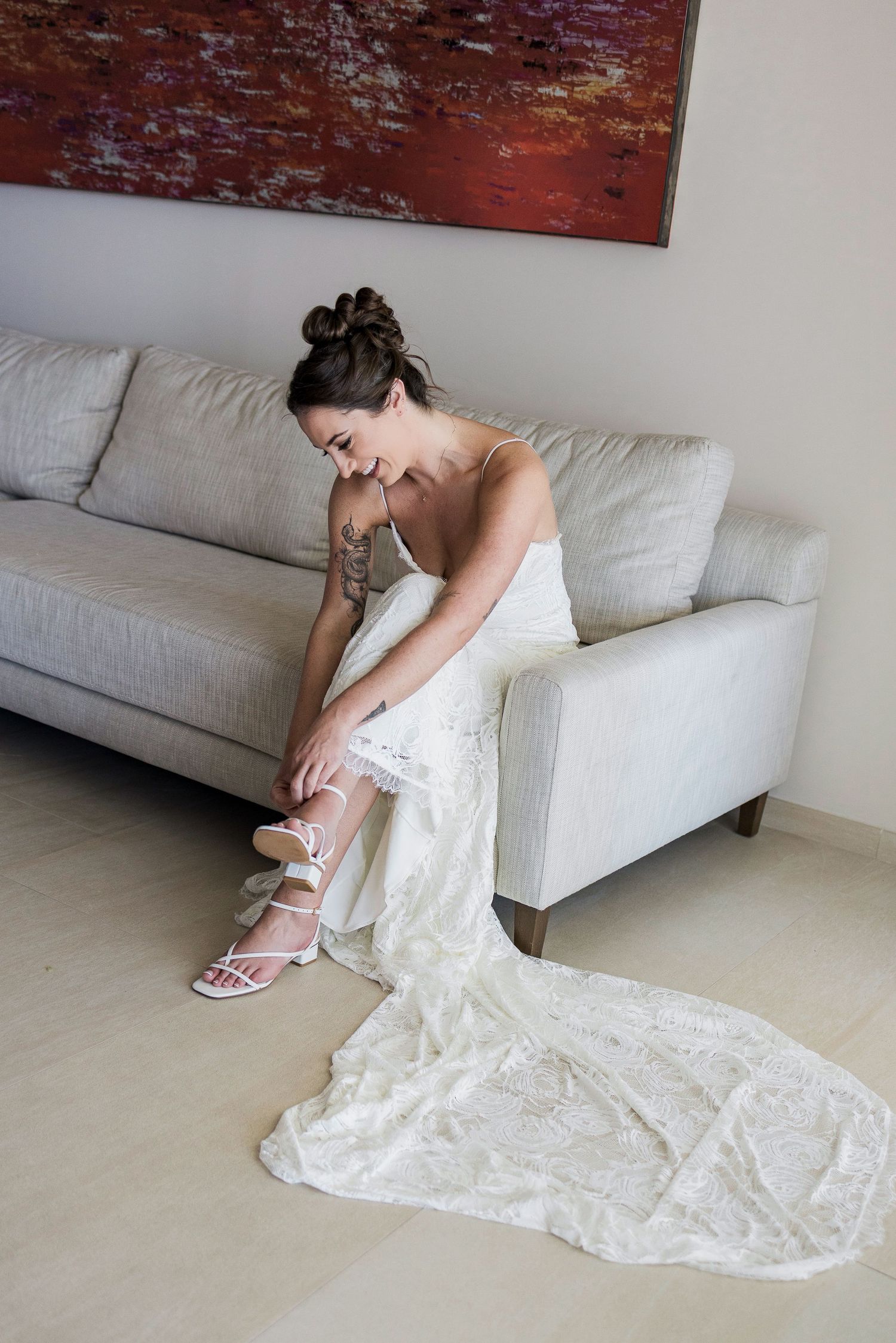 Bride in white lace gown sitting on gray sofa, fastening strappy heels, with abstract art on wall behind her.
