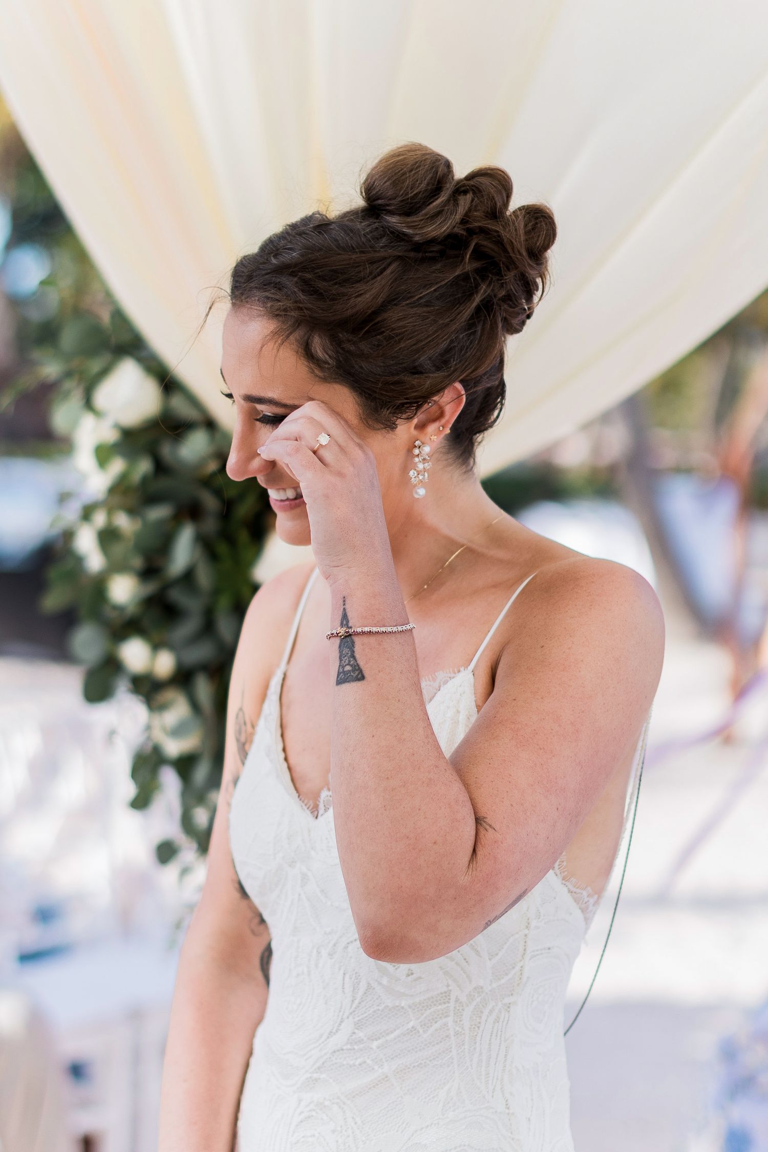 Emotional bride in white lace gown wiping tears at outdoor wedding ceremony with floral backdrop.
