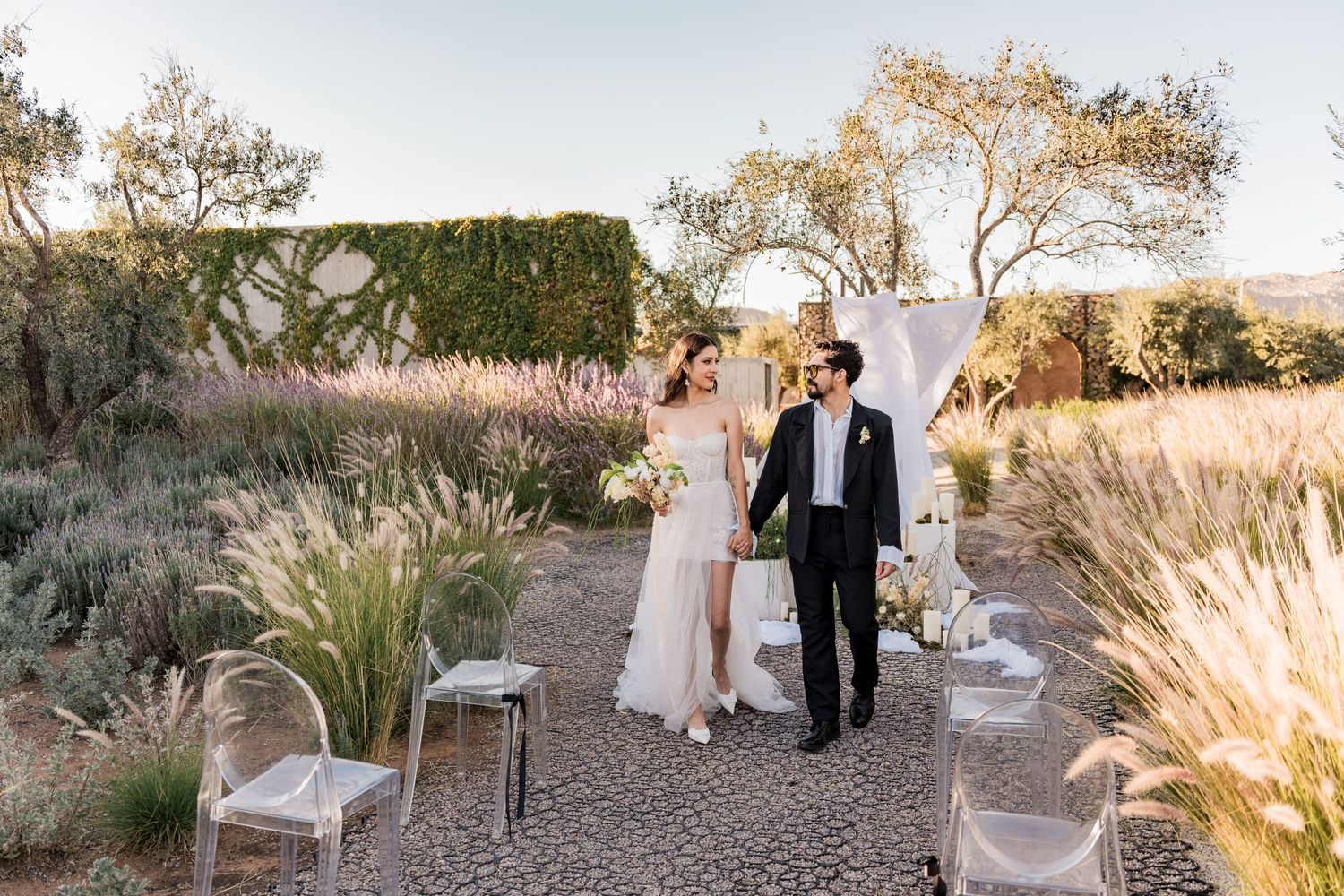 A bride and groom walk together through a stunning outdoor garden wedding ceremony at golden hour.