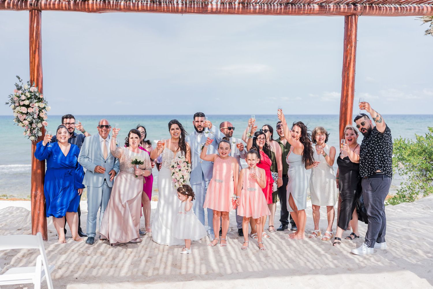 Wedding party celebrating with raised hands under a wooden pergola with ocean views behind them.