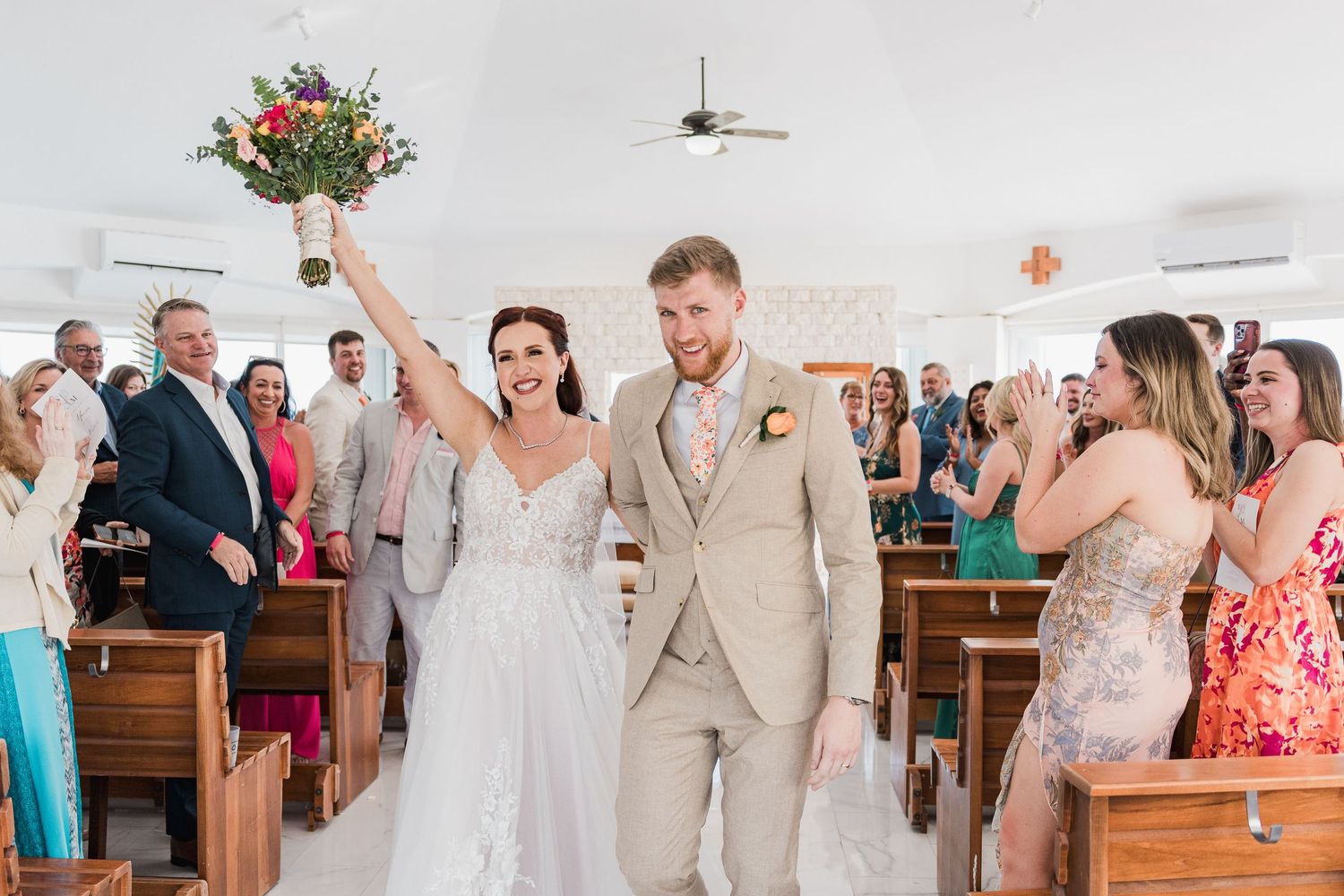 Joyful bride raises bouquet as couple exits white chapel after wedding ceremony with cheering guests.
