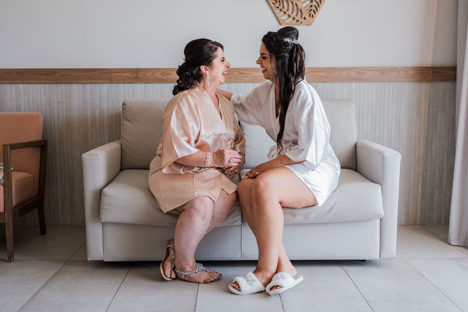 Bride and bridesmaid in robes sharing a joyful moment seated on a couch before wedding.