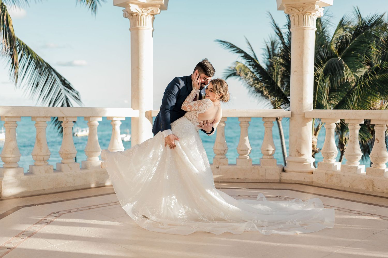 Groom dips bride on ocean-view terrace with palm trees and blue water backdrop.