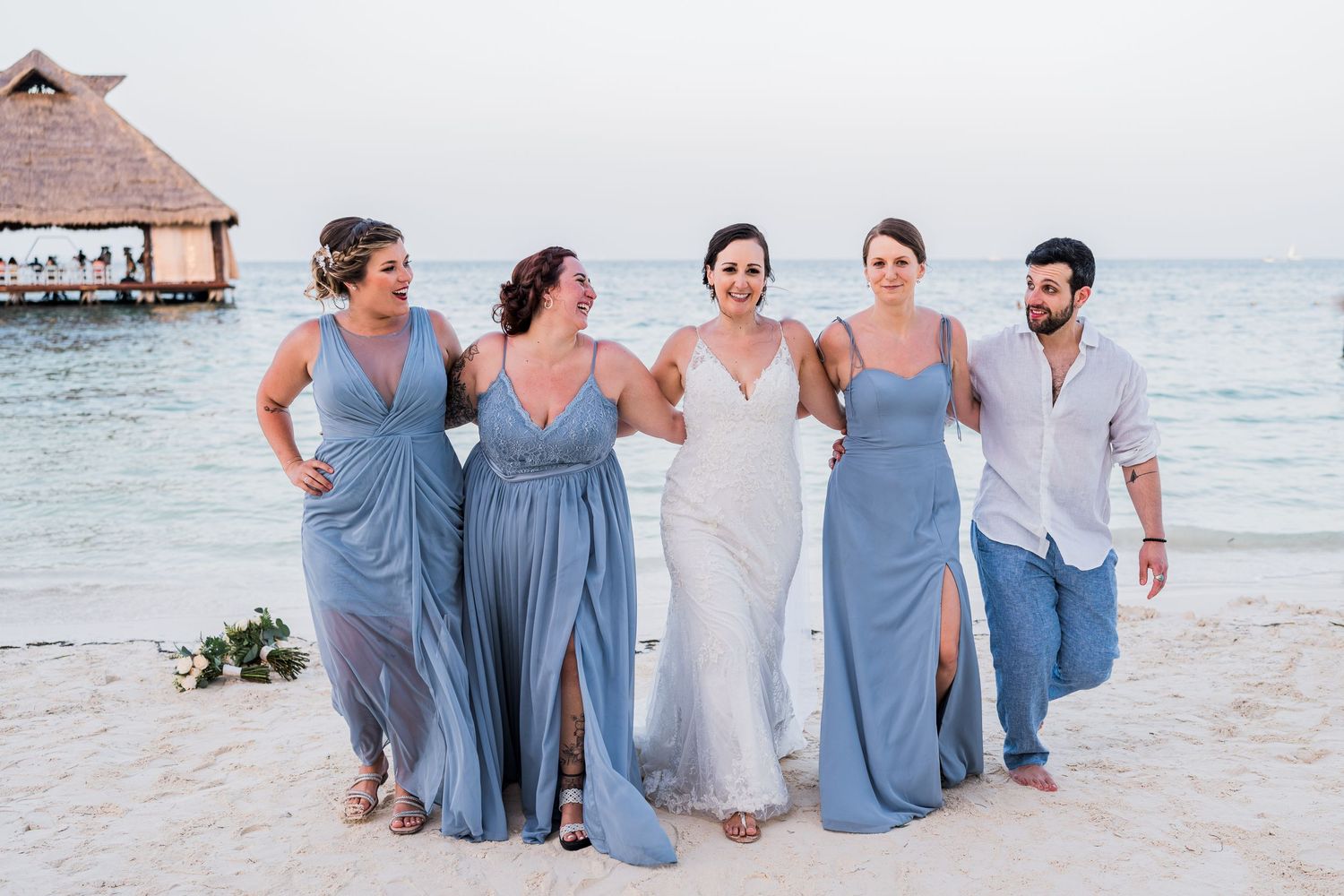 Bride walks with bridesmaids in blue dresses on a beach with ocean views at a destination wedding.