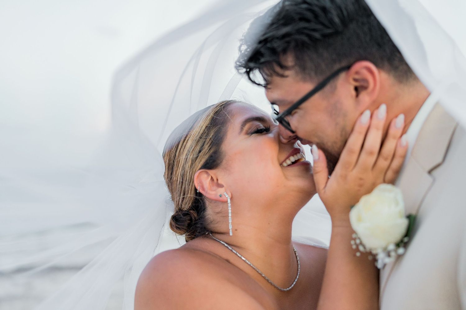 Bride and groom sharing a romantic kiss under a flowing white veil on their beach wedding day.