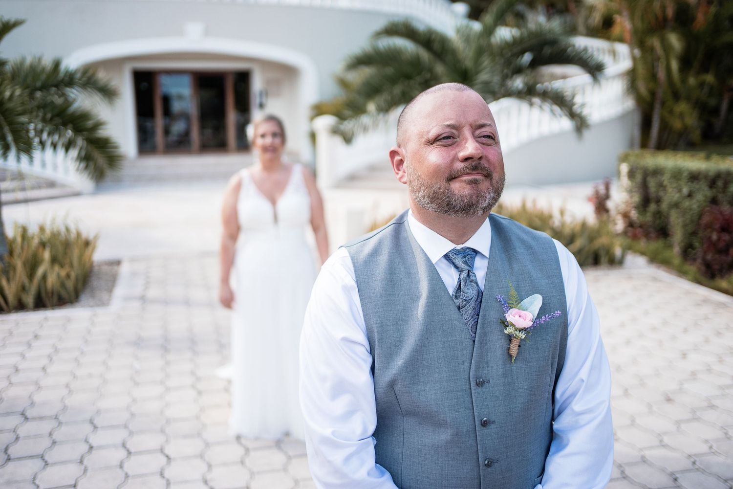 Emotional groom in gray vest with boutonniere awaits bride during outdoor first look ceremony.