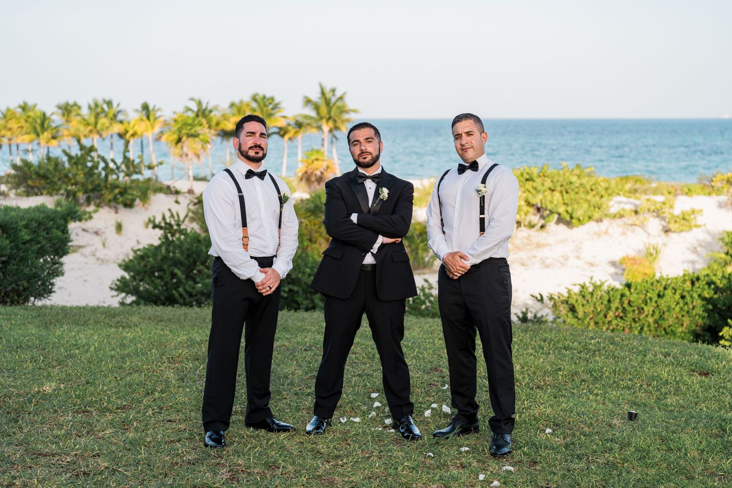 Three groomsmen in black tuxedos and suspenders pose seriously by a tropical beach.