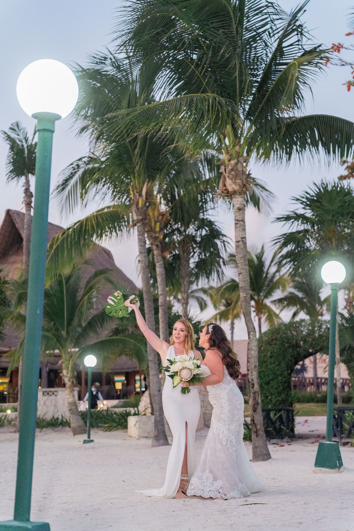 Two brides in white gowns celebrating on tropical beach with palm trees at dusk.