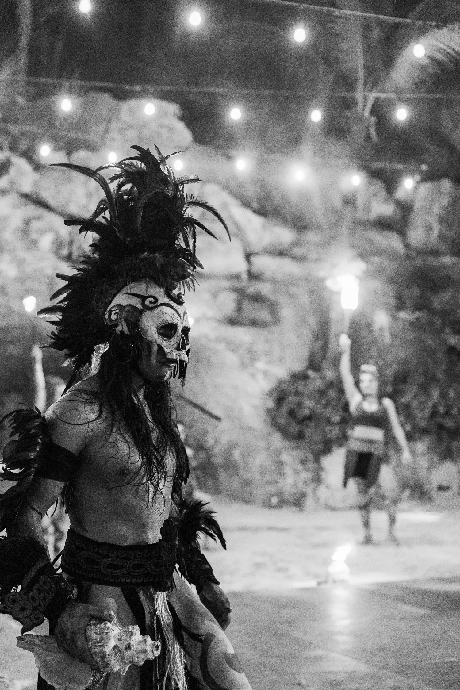Black and white photo of a performer in Mayan skull costume with feathered headdress at an outdoor night show.