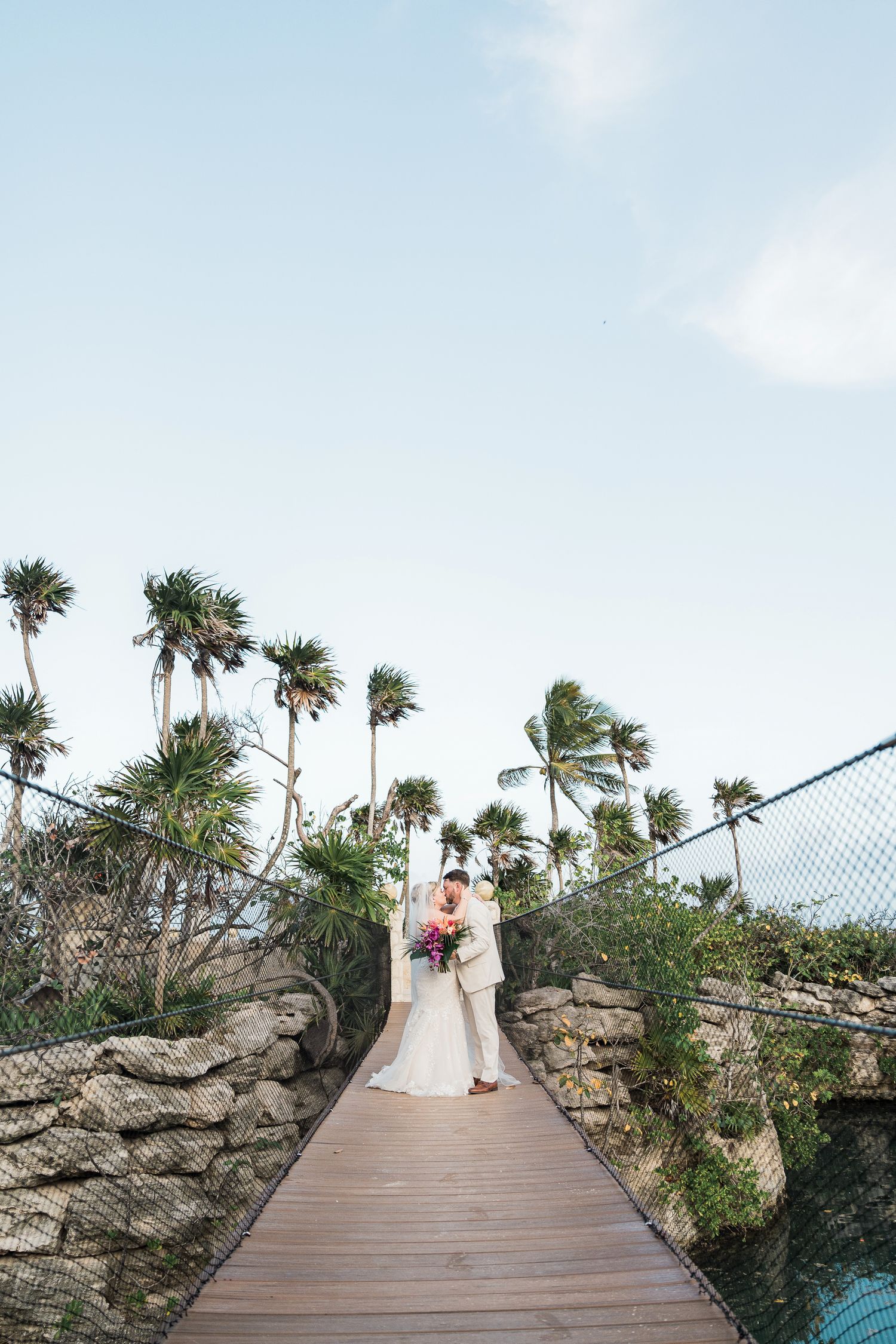 Hotel Xcaret México Wedding - Bride and groom embrace on a wooden suspension bridge surrounded by palm trees