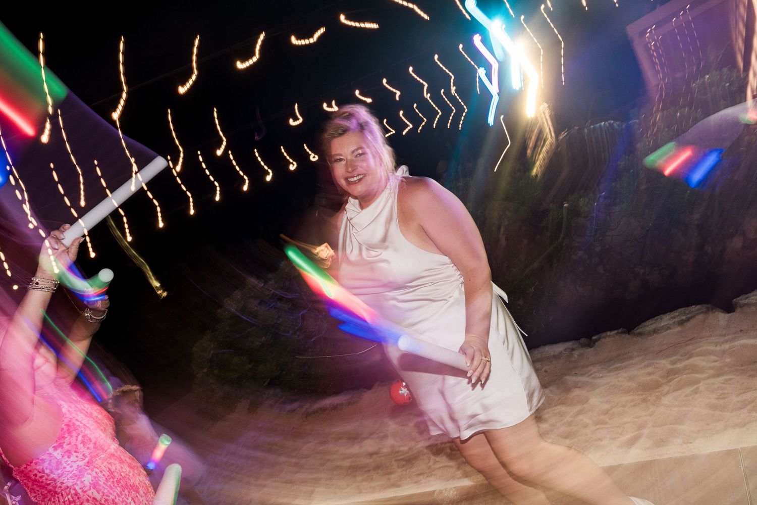 Hotel Xcaret México Woman in white dress dancing at night beach party with colorful glowing light trails and festive illuminations.