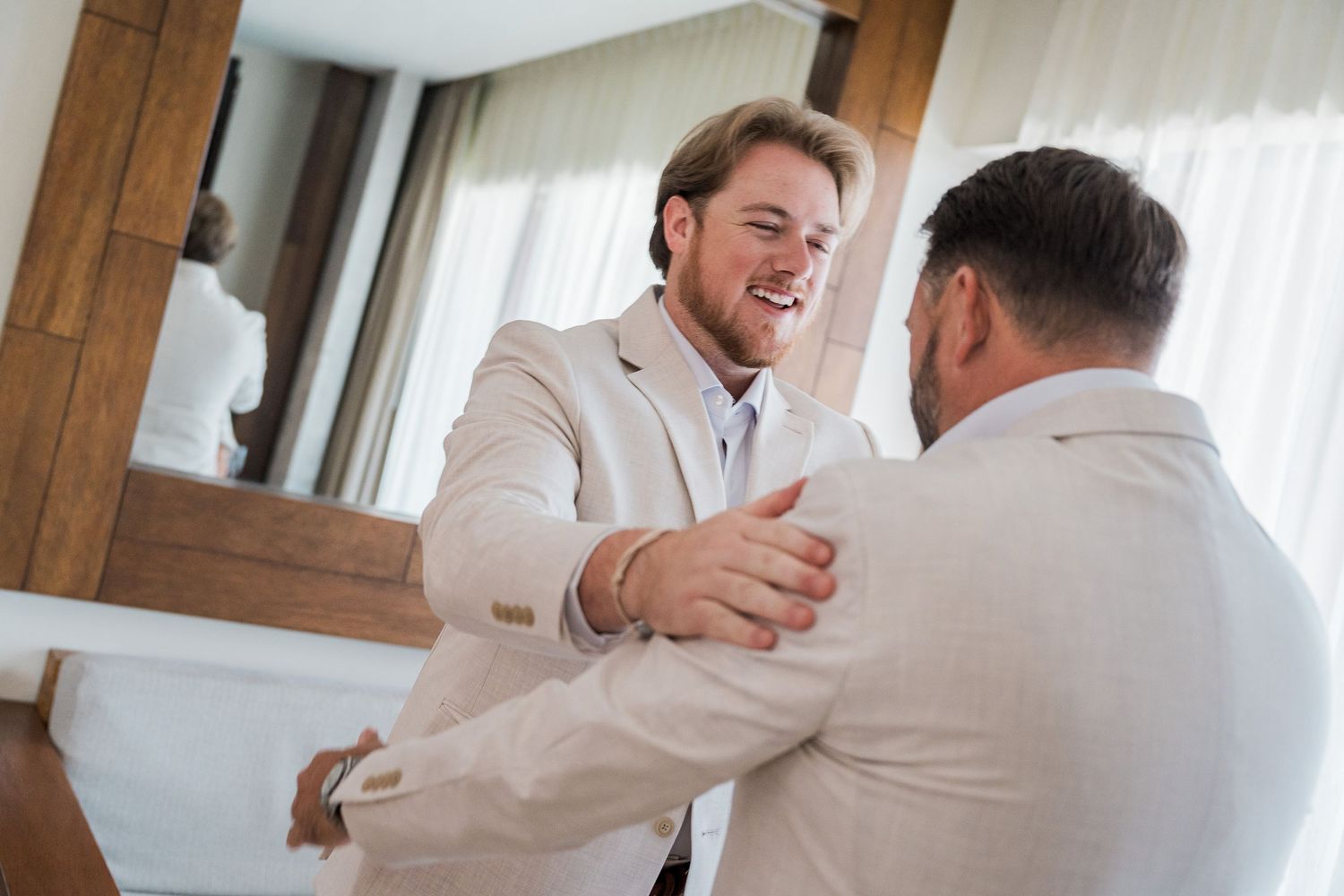 Two men in beige suits laughing and embracing at a wedding celebration indoors.