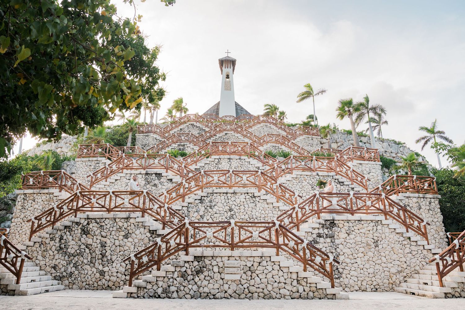 Stone pyramid structure with wooden staircases and a white spire, surrounded by tropical palm trees.