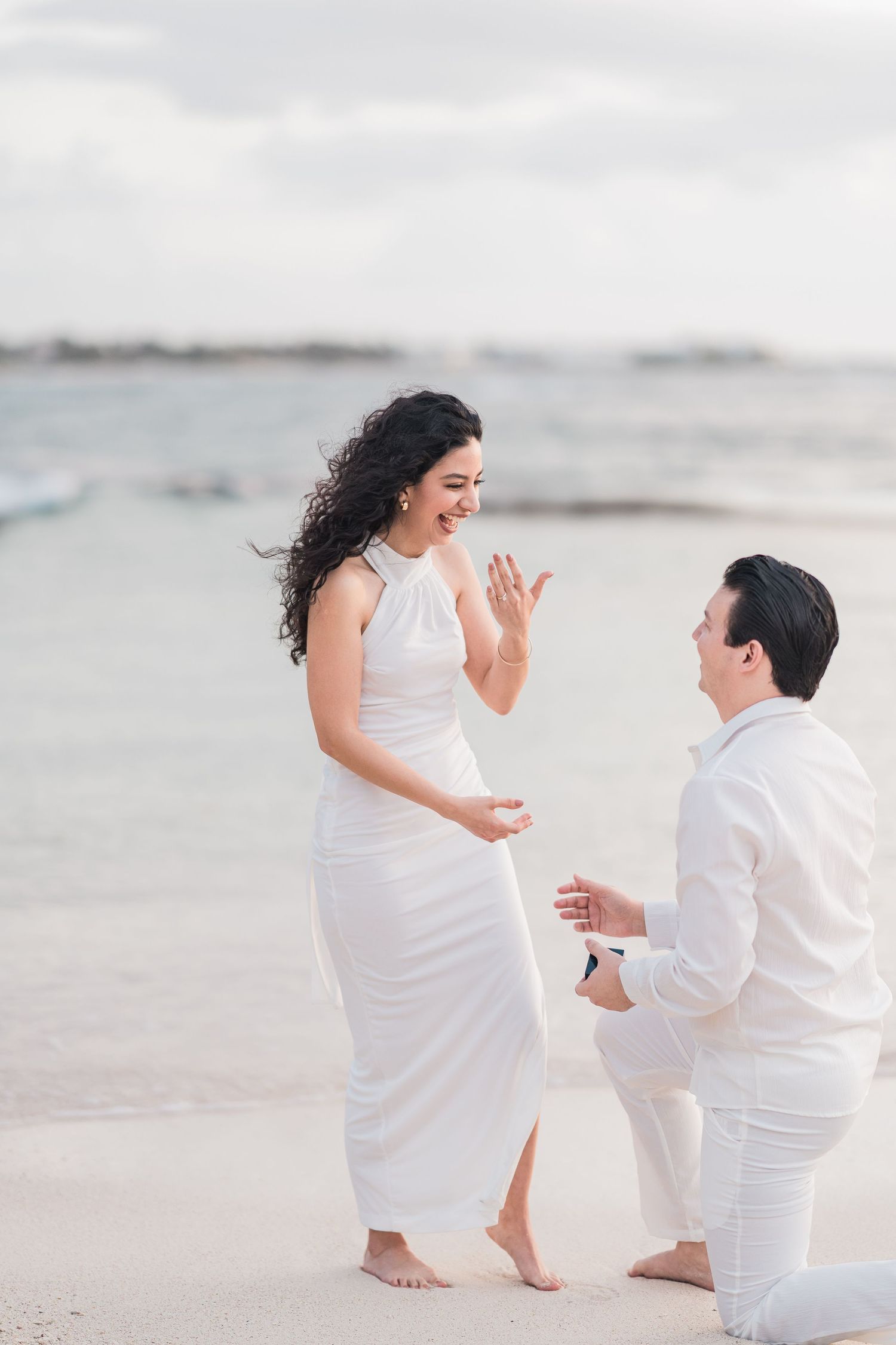 Man proposes on bended knee to woman in white dress on sandy beach, both dressed in white.