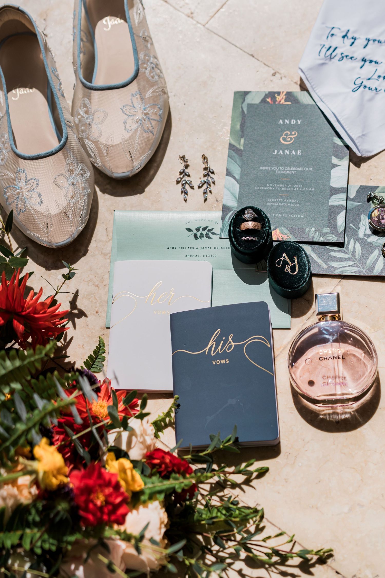 Wedding flat lay featuring vow books, Chanel perfume, floral invitations, bridal shoes, rings, and colorful flowers on marble.