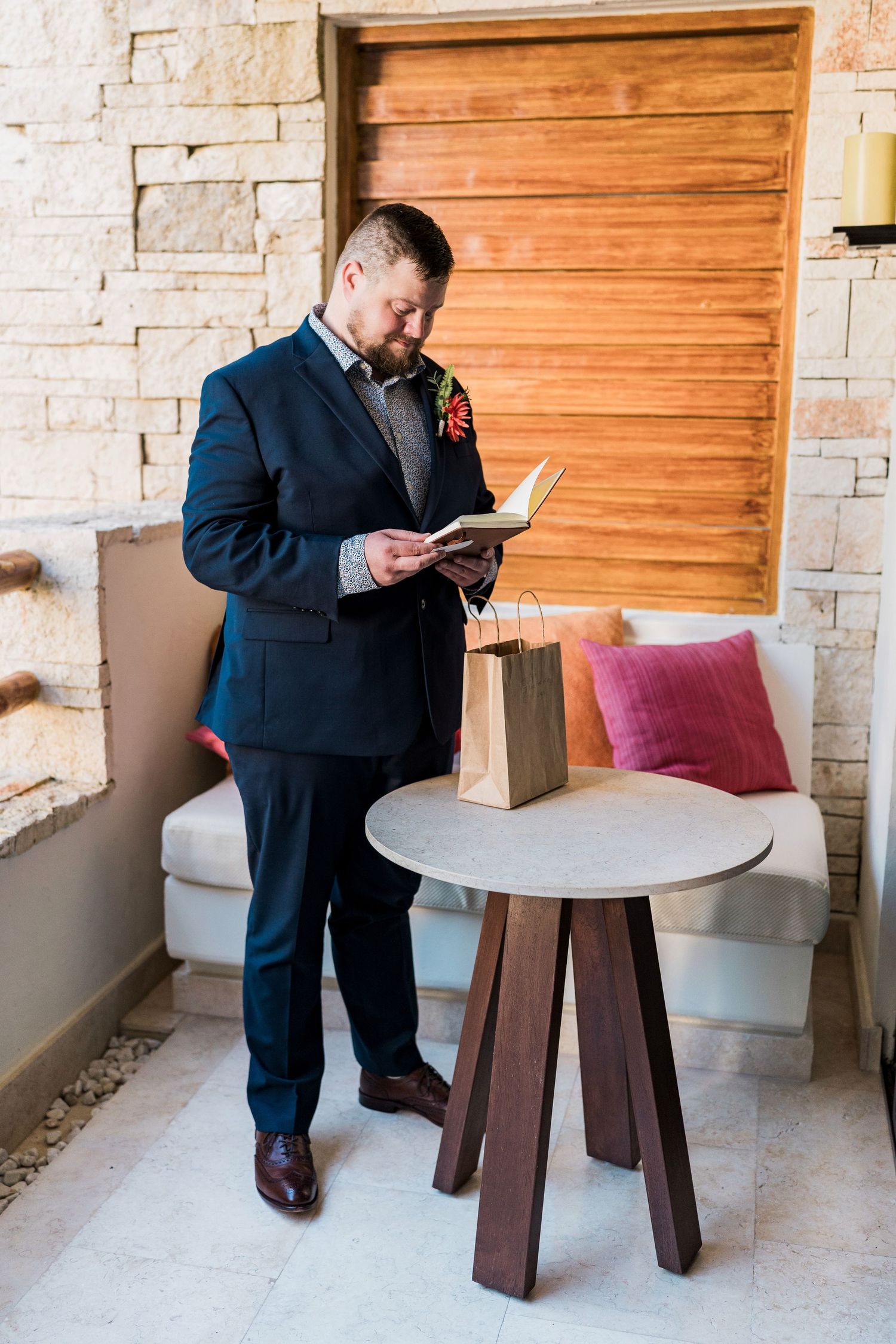 Secrets Akumal Wedding - Groom in navy suit reads a book beside a round wooden table with a gift bag in an elegant stone-walled venue.